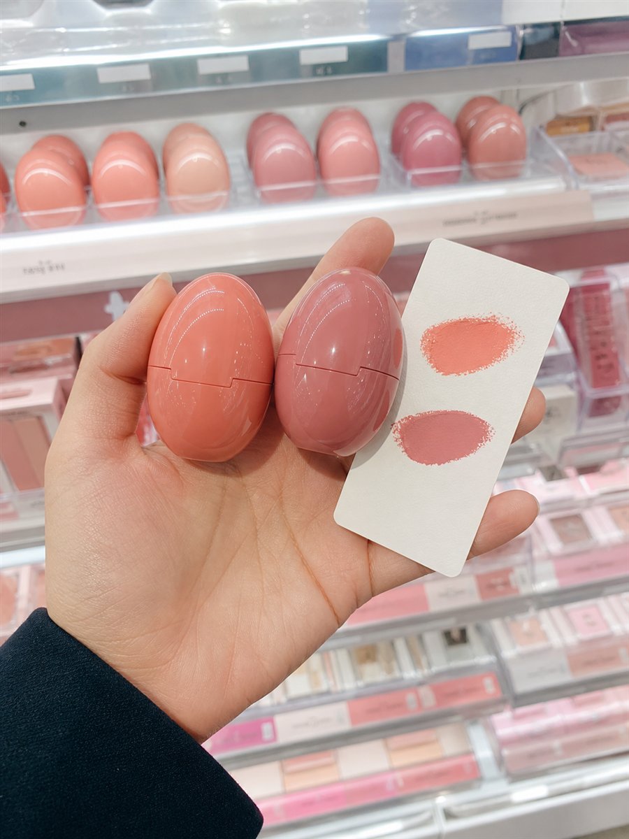 AI-assisted editorial visualization of generic egg-shaped cream-blush compacts and a disposable shade card in a Korean beauty store · package reference