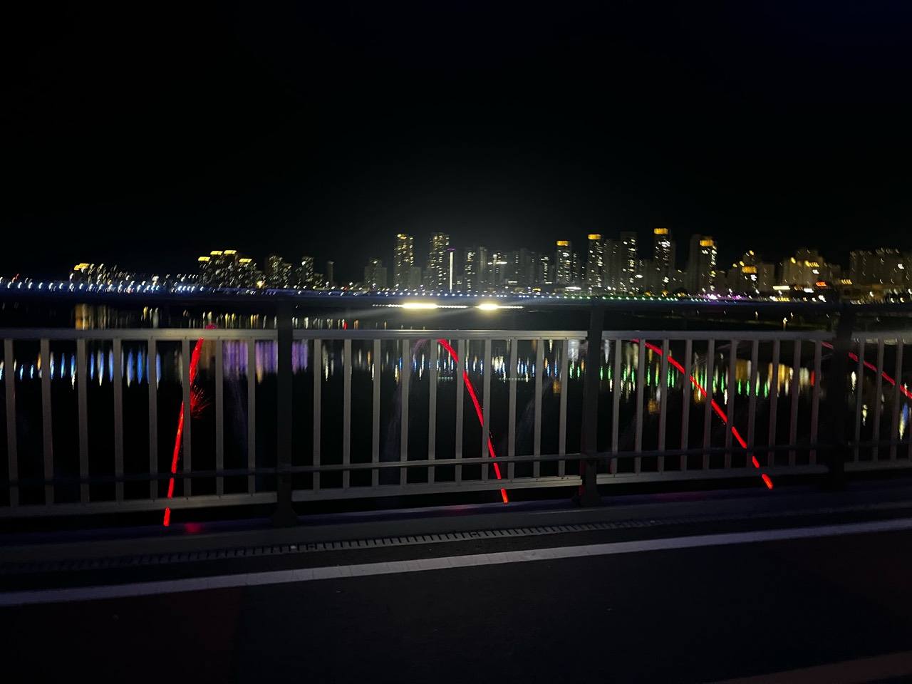 난간 사이 붉은 조명의 물줄기와 강 건너 아파트 야경